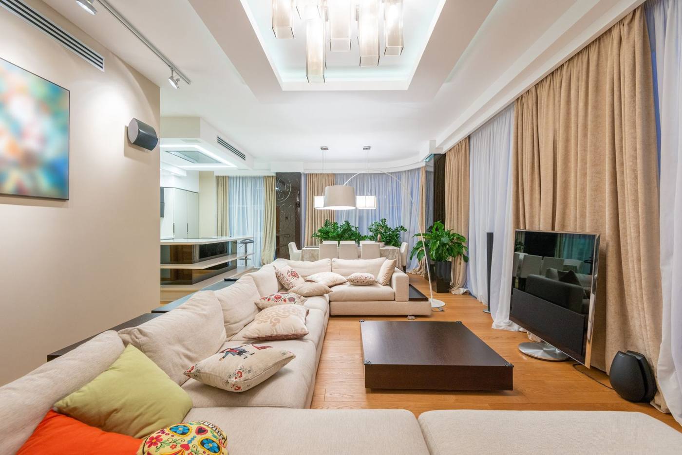 Living cu canapea bej, deschis spre bucătărie și zona de masă, într-un apartament luminat seara